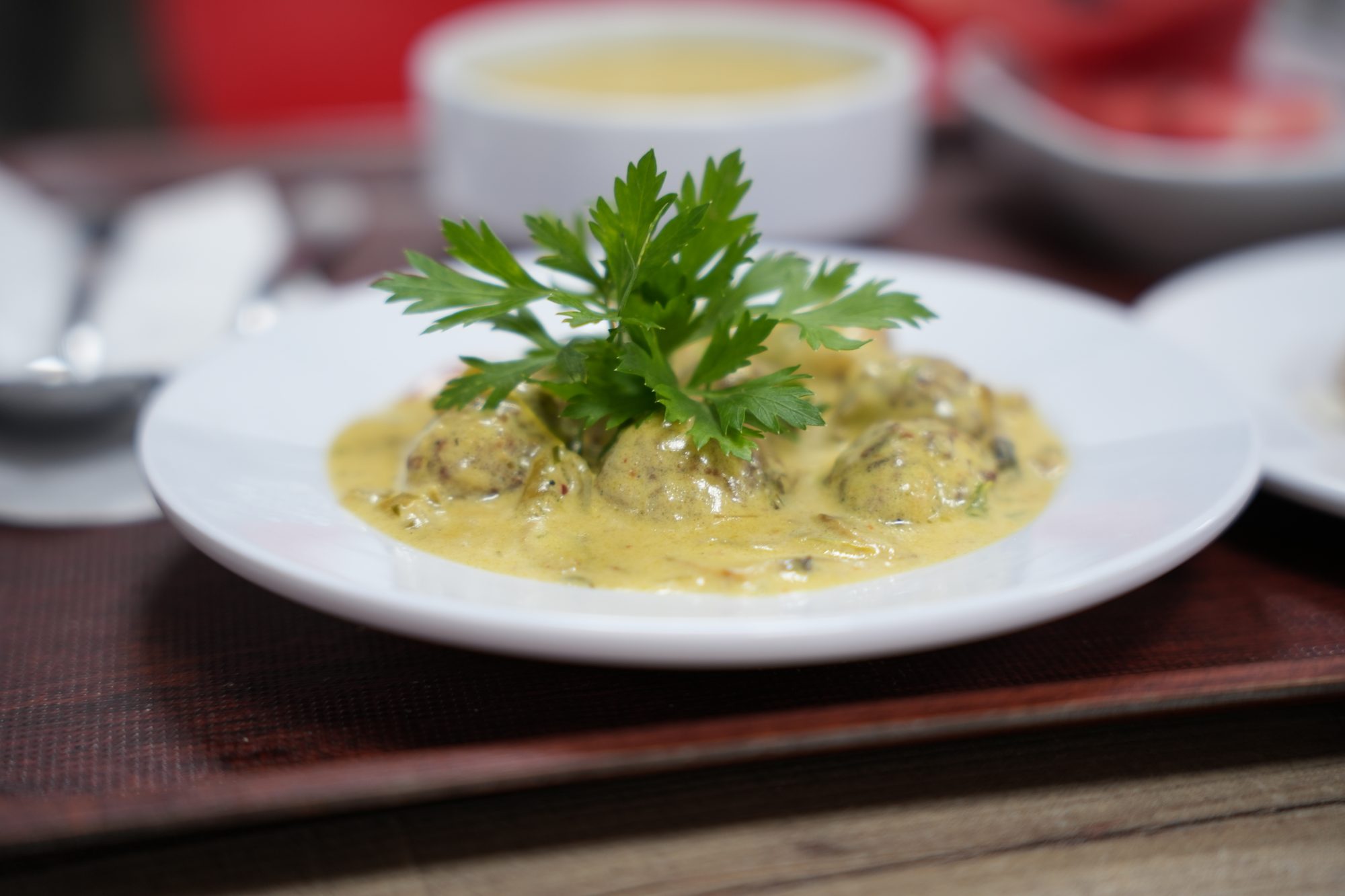 Okul yemekhanesinden bir tabak: yeşillikle süslenmiş soslu köfte; arkada çorba kâsesi.