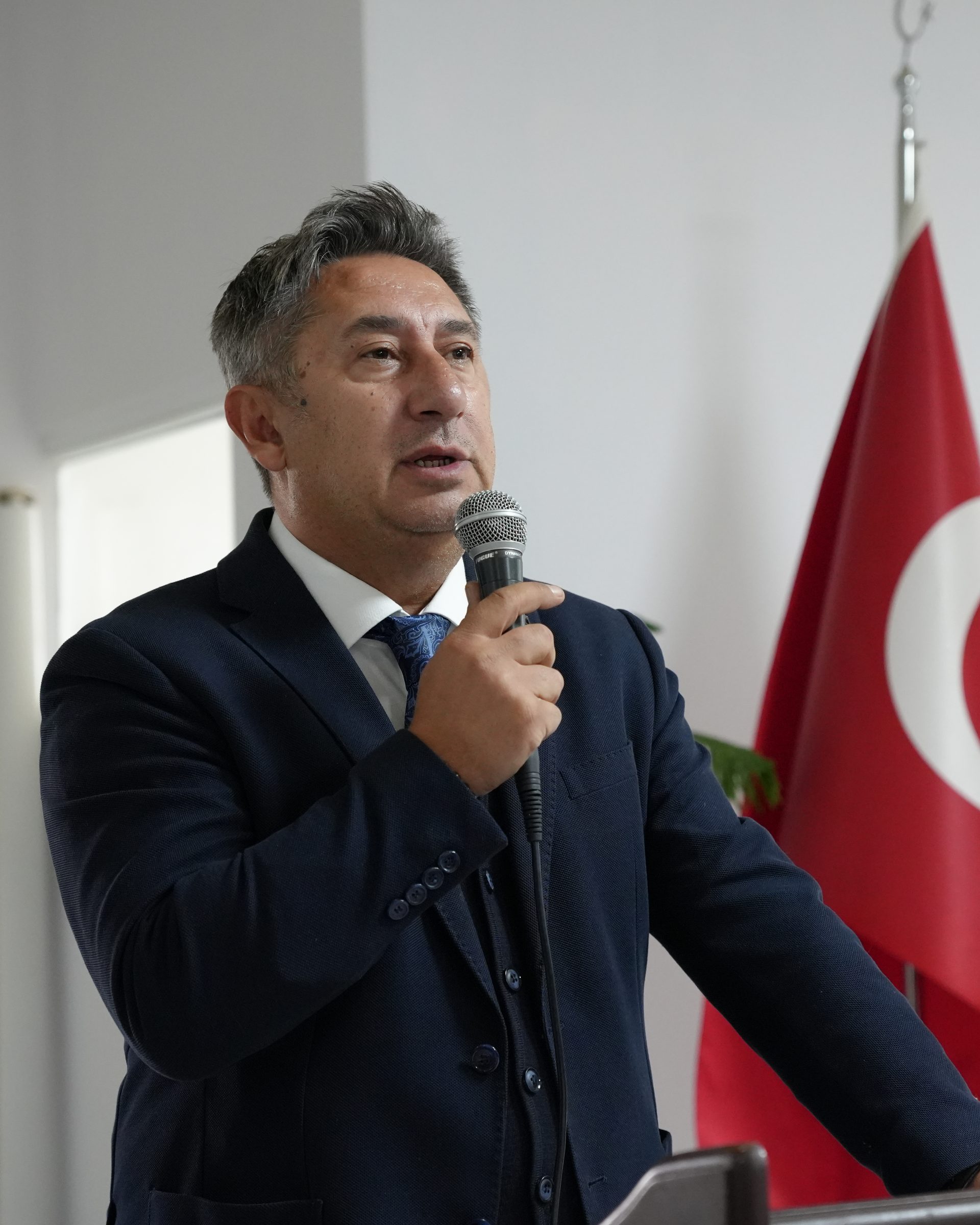 Öztuna Norman, lacivert takım elbiseyle kürsüde mikrofonla konuşuyor; arkasında Türk bayrağı.